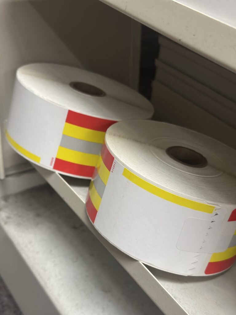 Rolls of red, yellow, and white direct thermal labels stacked on a shelf.